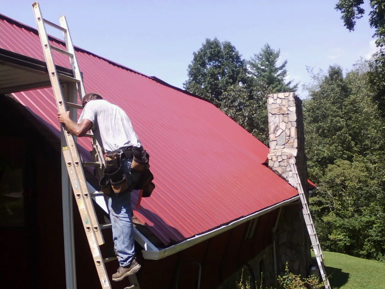 Roofer on a ladder beside a red metal roof and stone chimney — Soffit & Fascia Repair by Roofing Brothers in Berkeley